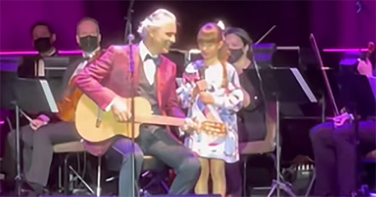 mężczyzna siedzi z gitarą, obok niego stoi dziewczynka, a w tle siedzi orkiestra