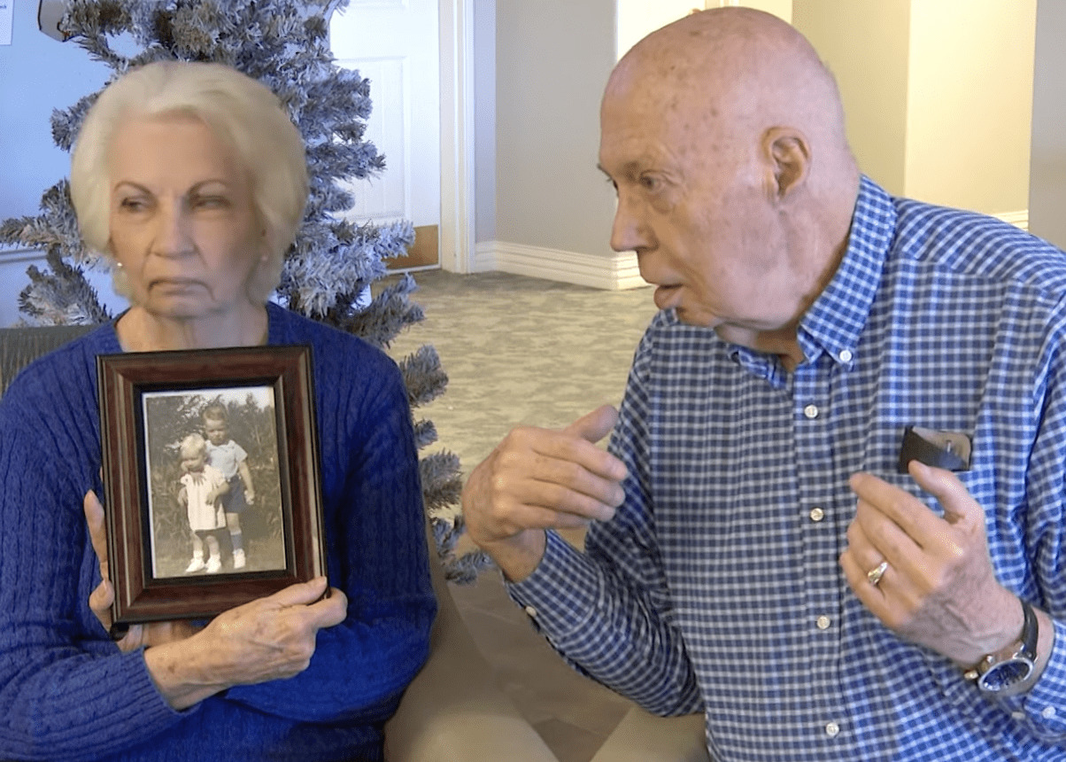 starsza kobieta trzyma w ręku ramkę ze zdjęciem, a starszy mężczyzna siedzi koło niej